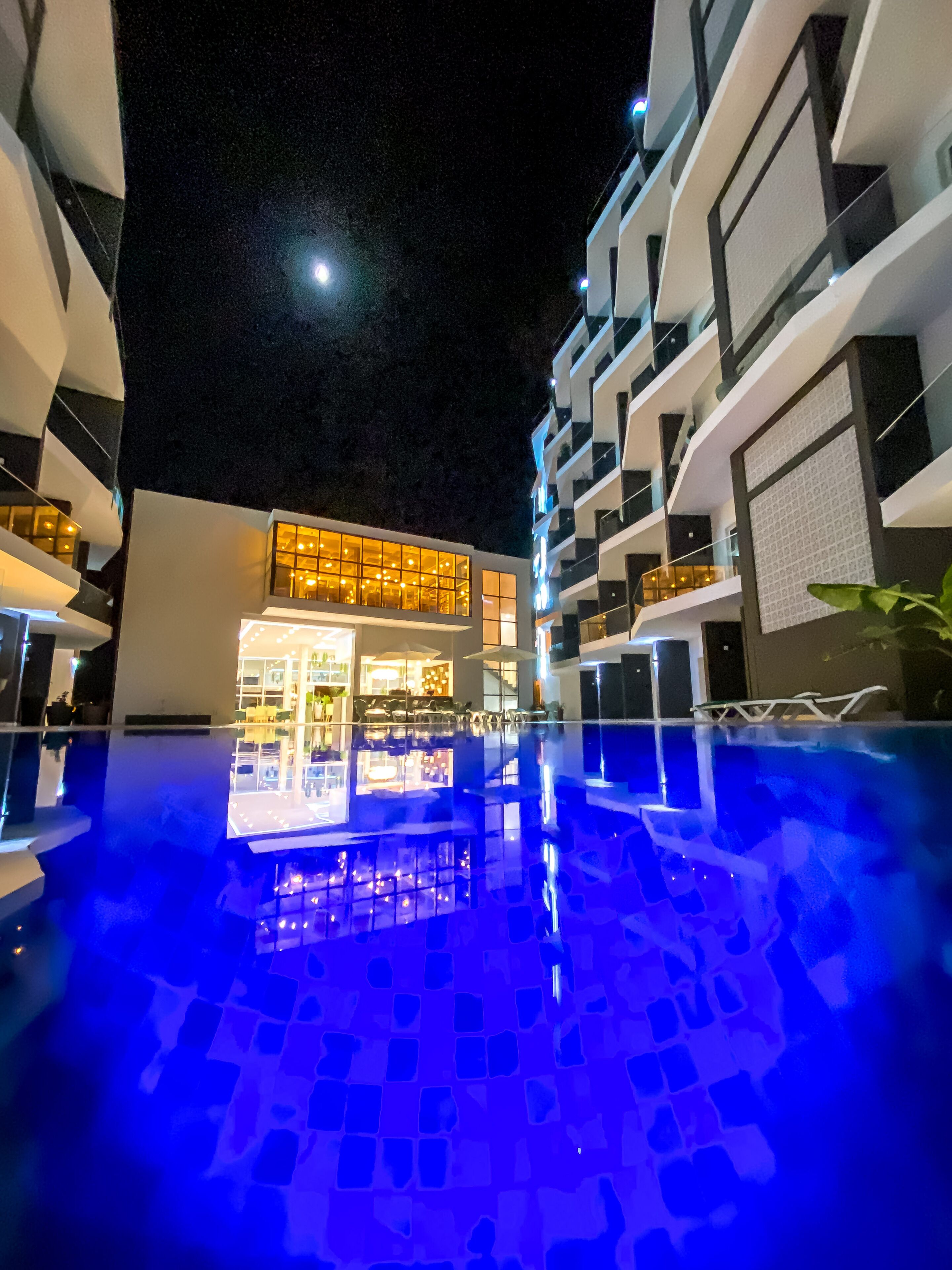 A modern hotel with a pool area, featuring a well-lit bar and lounge area, and a clear view of the pool's edge.