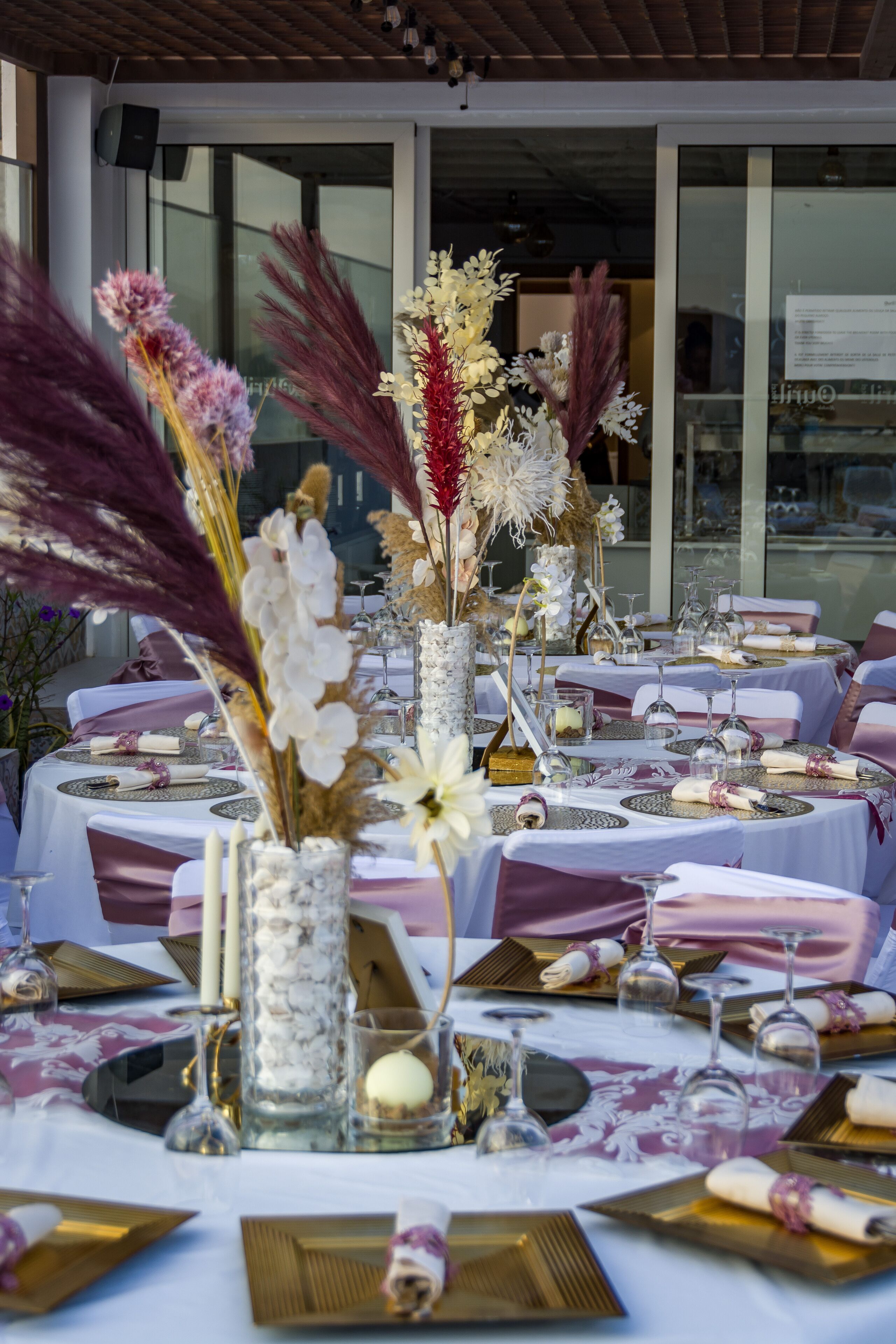 Une table dressée pour un événement formel, avec de la vaisselle blanche et dorée, des centres de table floraux et une structure en verre qui l’entoure.