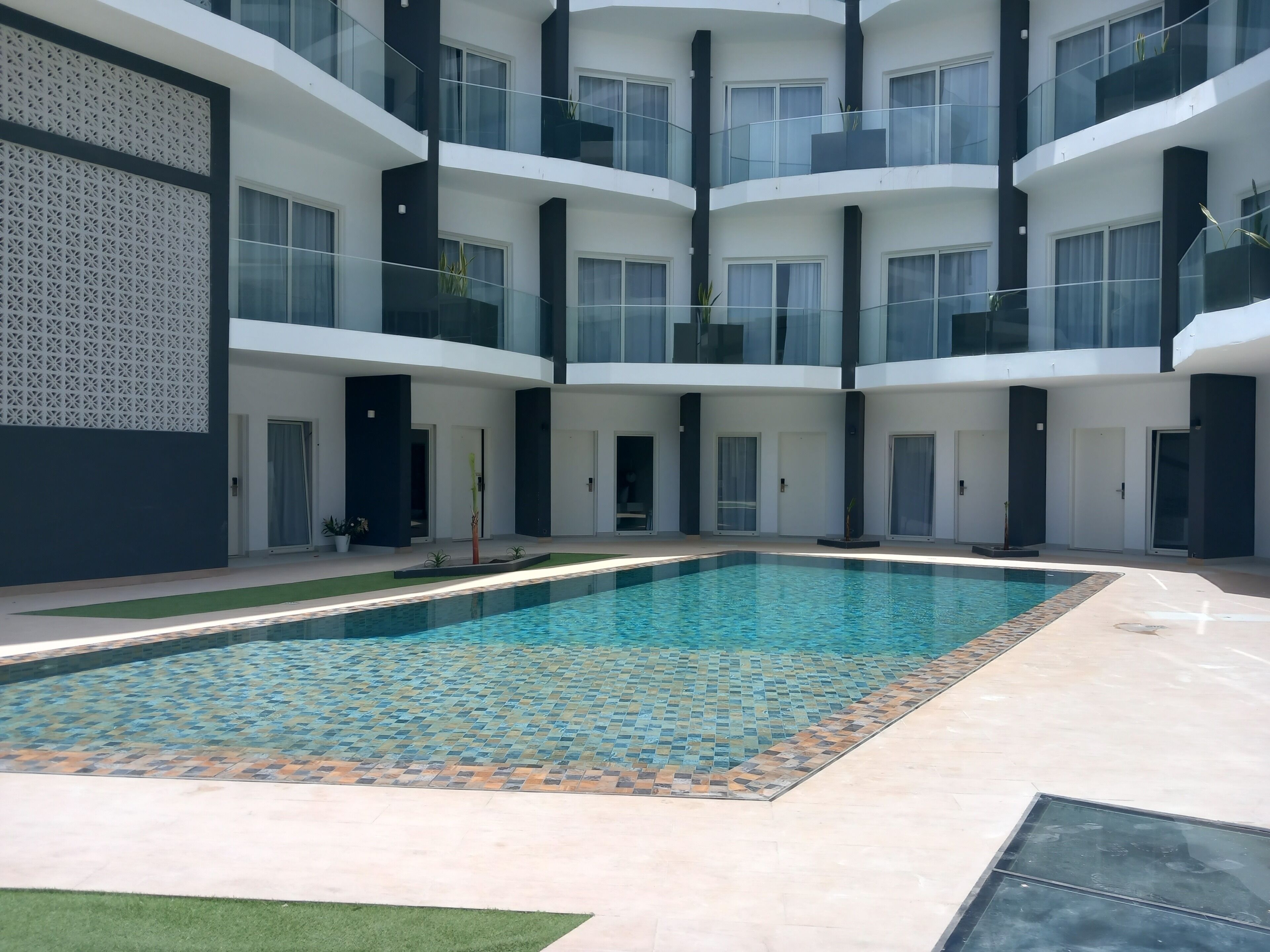 Un immeuble moderne à plusieurs étages, doté d’une piscine centrale, de balcons en verre et d’une terrasse autour de la piscine au revêtement décoratif.