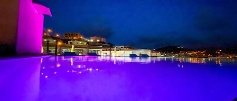 Vista noturna de uma piscina com borda iluminada em roxo, refletindo edifícios e luzes próximos.