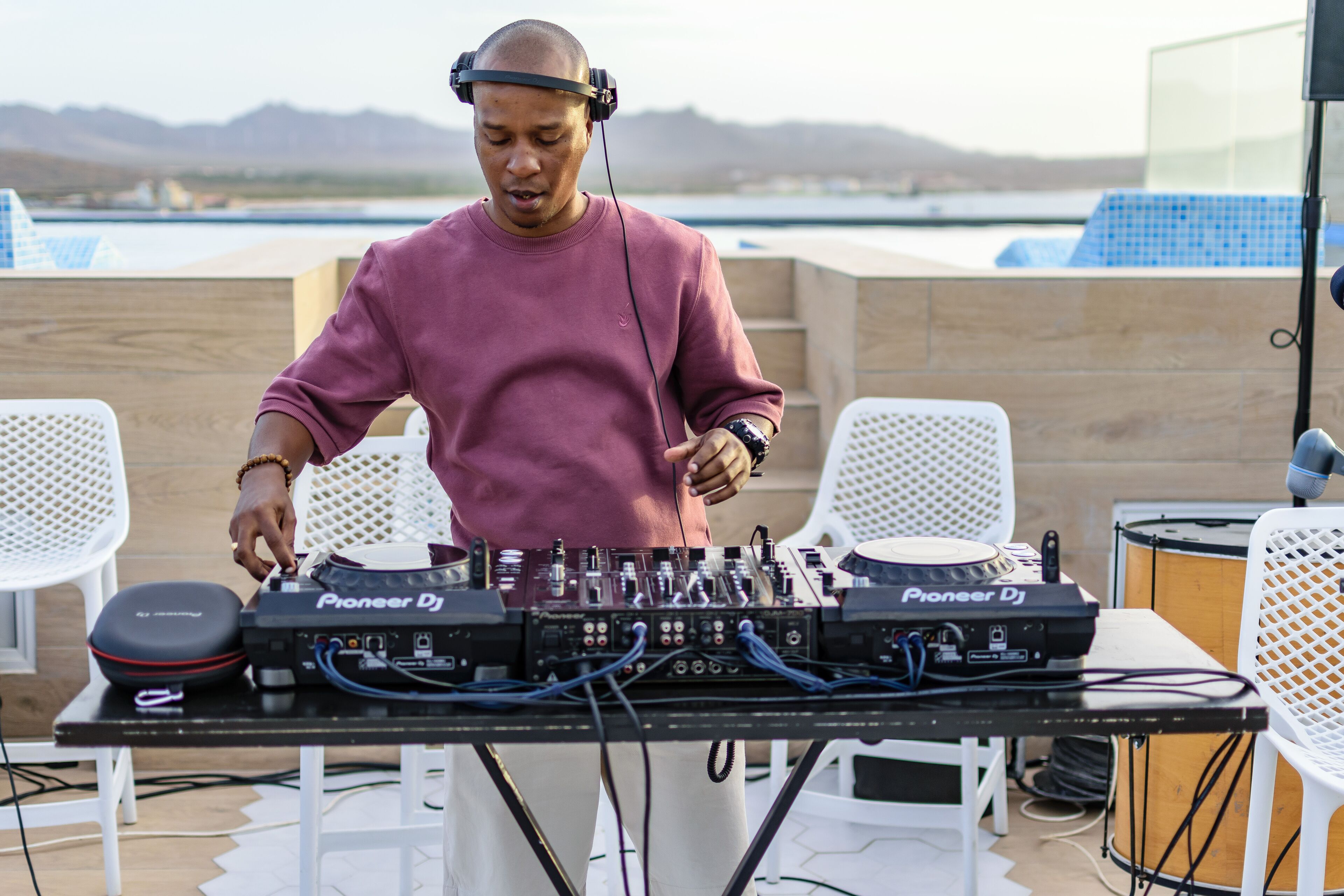 A DJ setup with Pioneer DJ equipment on an outdoor table, surrounded by white chairs, with a scenic view in the background.