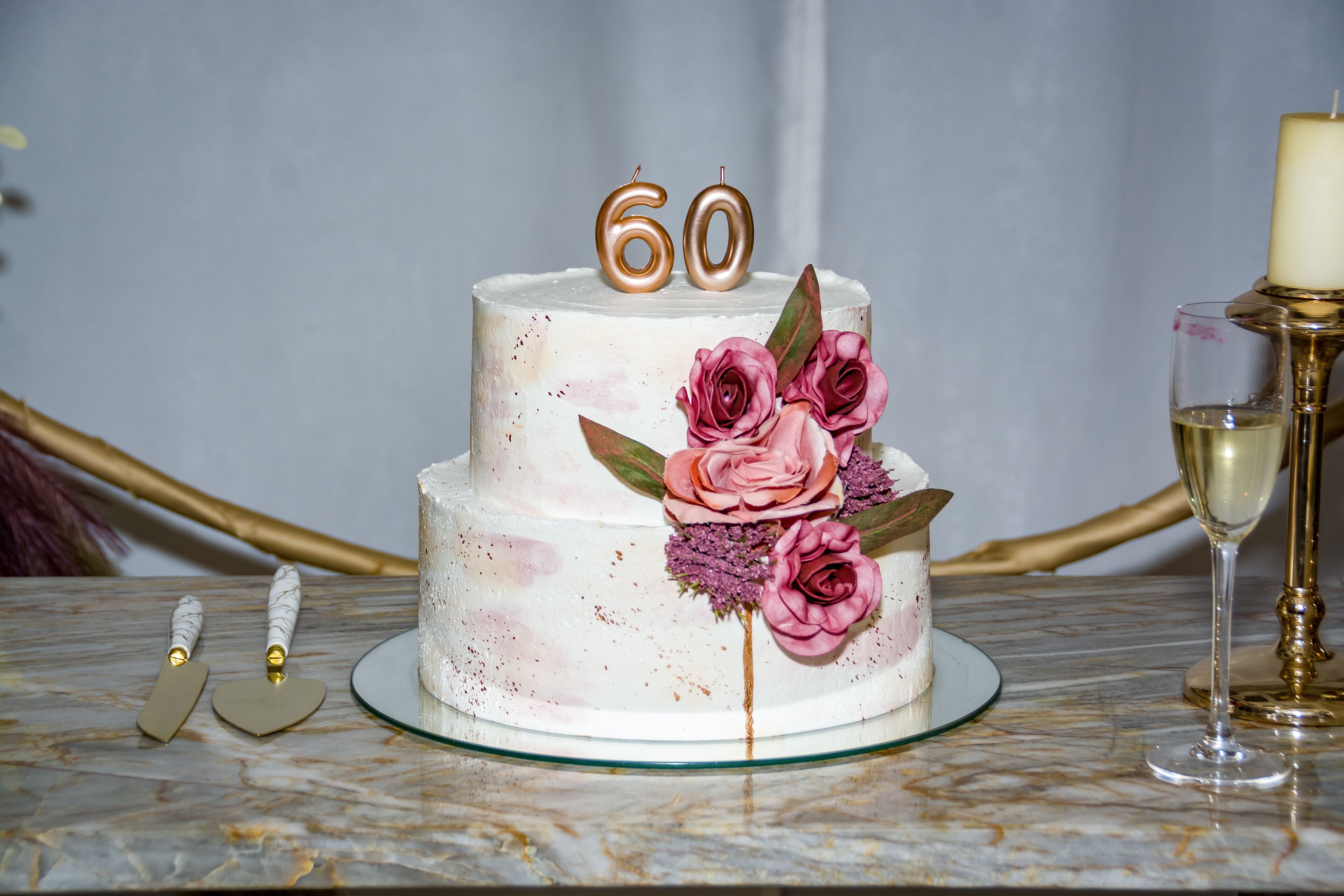 A two-tiered cake with '60' candles on top, decorated with pink roses and leaves, placed on a marble table.