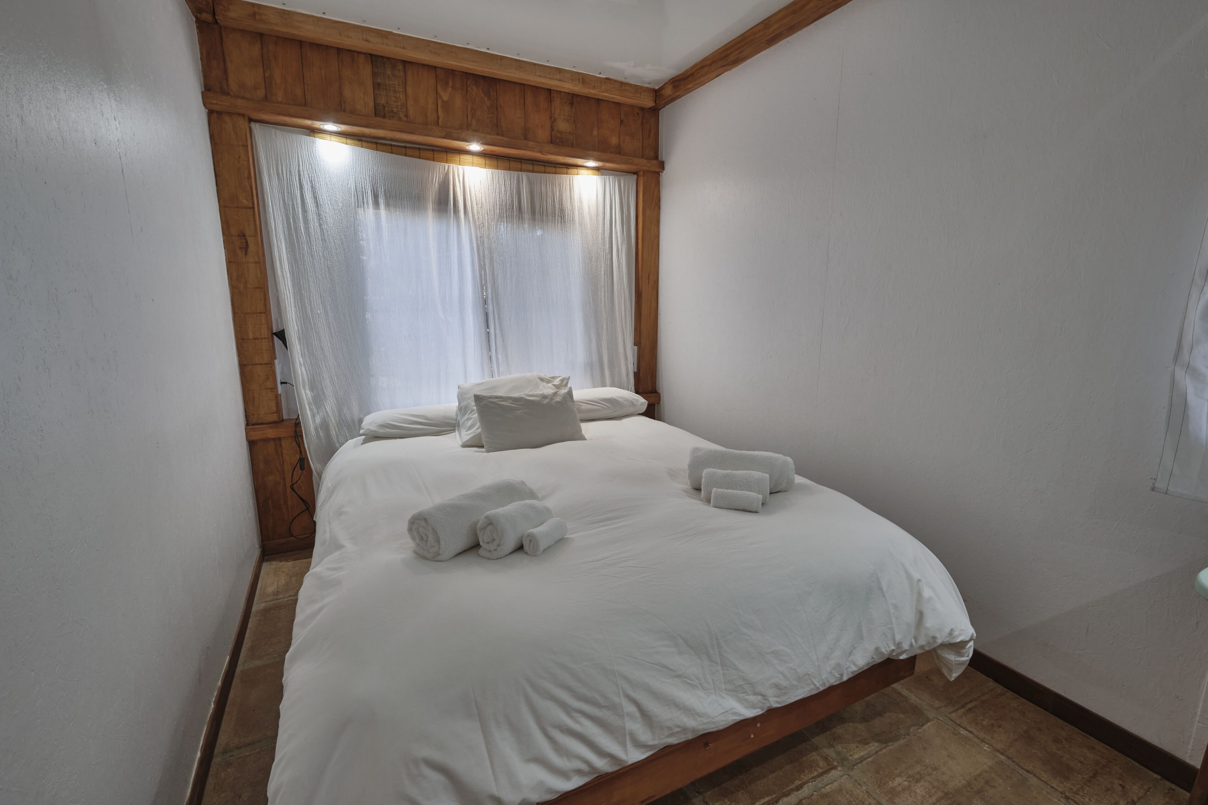 Ein ordentlich bezogenes Bett mit weißen Leinen und Handtüchern, ein Holz-Kopfteil und ein Fenster mit durchsichtigen Vorhängen.