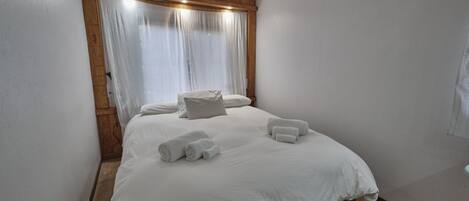 Ein ordentlich bezogenes Bett mit weißen Leinen und Handtüchern, ein Holz-Kopfteil und ein Fenster mit durchsichtigen Vorhängen.