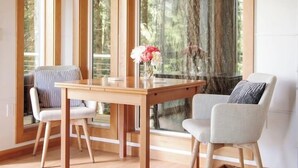 Una sala de estar moderna con una mesa de comedor de madera, dos sillas, un sofá y un ventanal con vistas a árboles.
