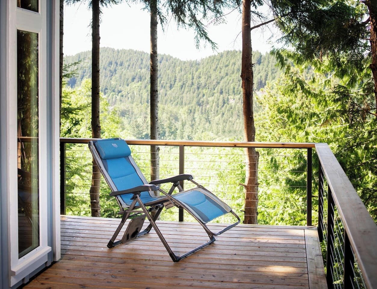 A wooden deck with two blue lounge chairs, overlooking a forested hillside.