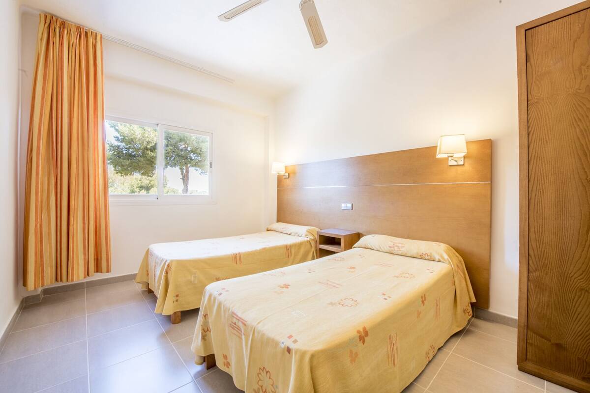 Una habitación de hotel con dos camas individuales, cabecero de madera, mesita de noche, ventana con cortinas y ventilador de techo.