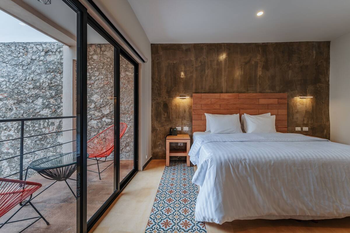 A bedroom with a large bed, a wooden headboard, two bedside tables with lamps, a patterned rug, and a balcony with chairs.