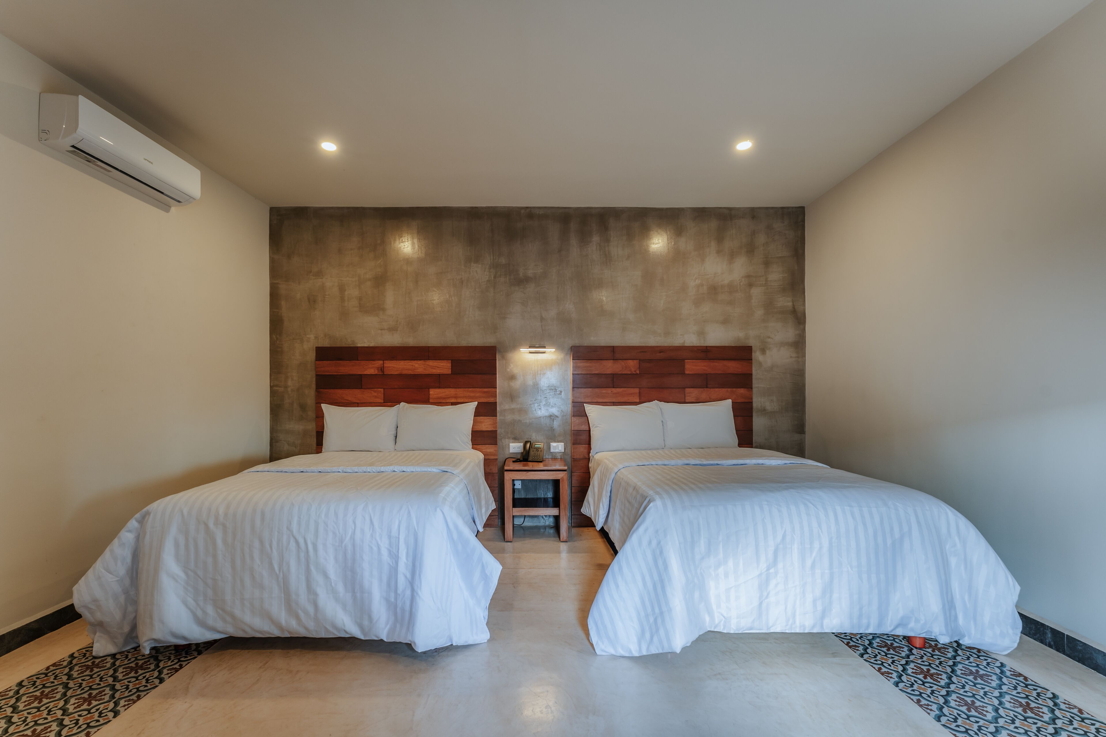 Quarto de hotel com duas camas, uma cabeceira de madeira, uma mesinha com abajur e um ar condicionado.