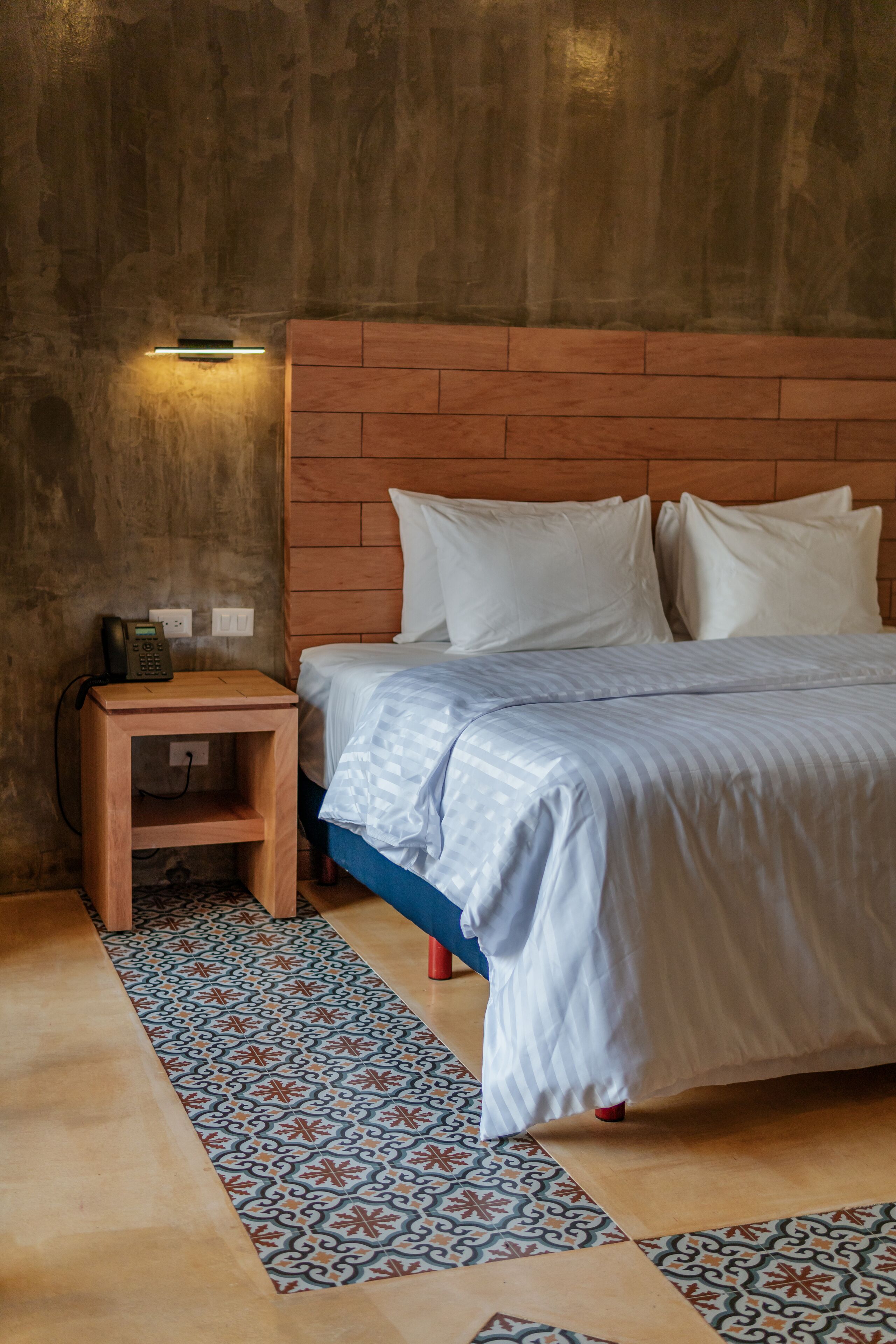 Ein Schlafzimmer mit einem Holzkopfteil, einem Nachttisch mit Telefon und einem Bodenbelag aus gemusterten Fliesen.
