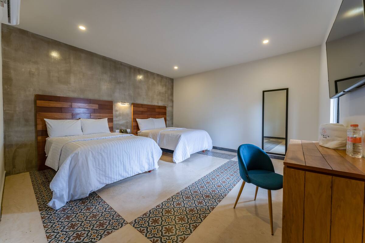 A modern hotel room with two beds, a wooden headboard, a patterned rug, a blue chair, and a desk with a mirror and a bottle.