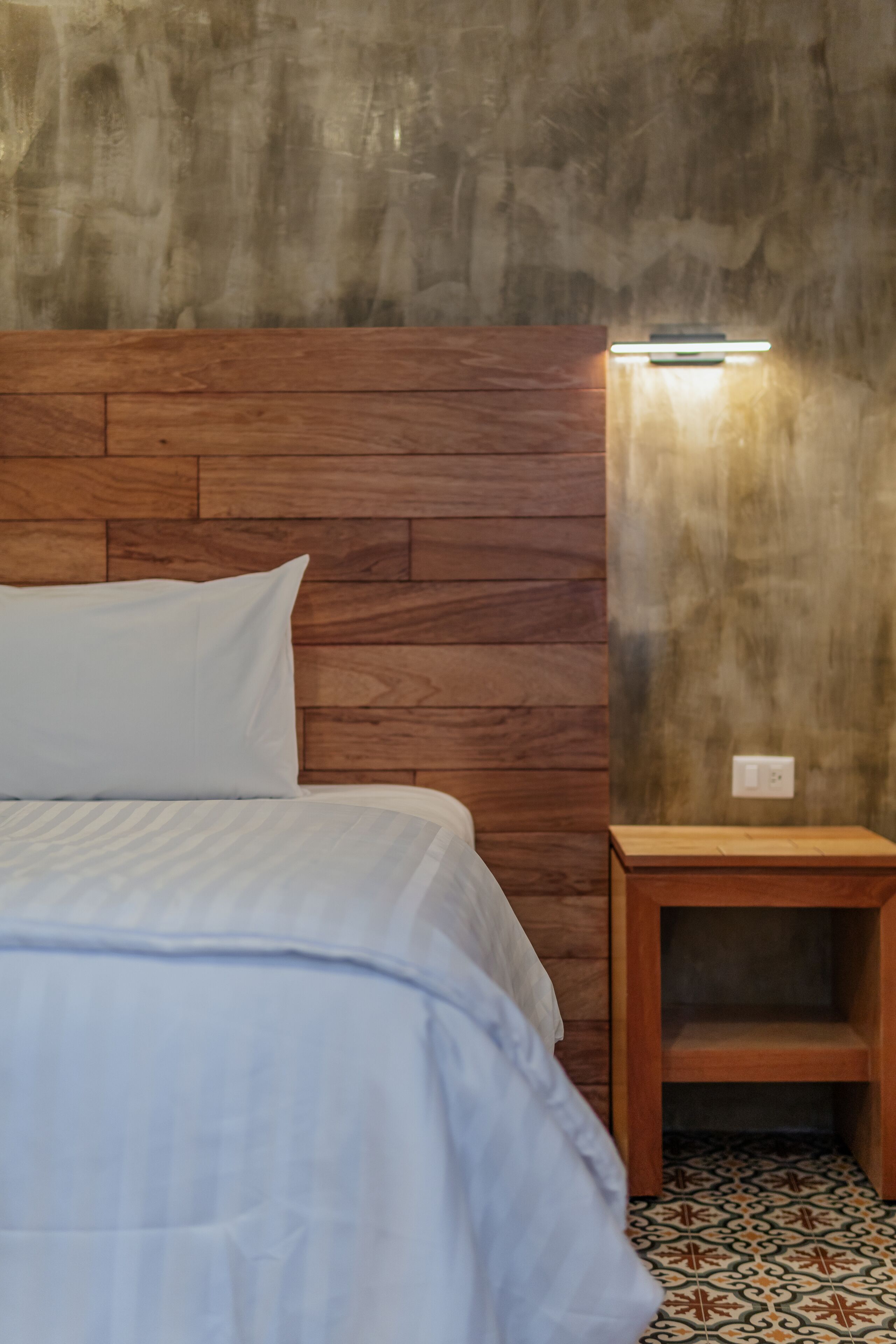 A bedroom with a wooden headboard, a bedside table, and patterned floor tiles.