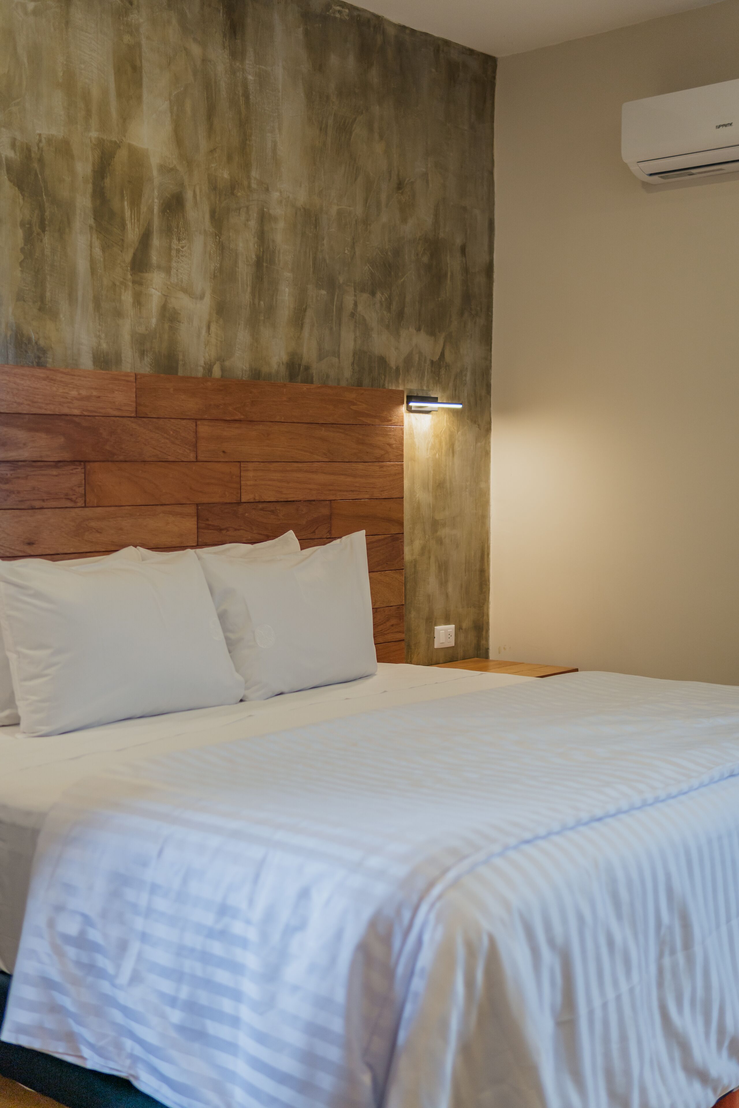 A neatly made bed with white linens and pillows, a wooden headboard, and a wall-mounted air conditioner.