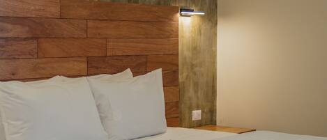 A neatly made bed with white linens and pillows, a wooden headboard, and a wall-mounted air conditioner.