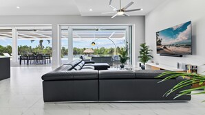Ein modernes Wohnzimmer mit einer großen Eckcouch, einem Deckenventilator und Blick auf einen Pool und eine Außenlounge.