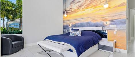 A modern bedroom with a large bed, a view of the outdoors through a window, and a sunset mural on the wall.