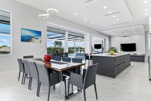 A modern kitchen with a central island, a dining area with a large table and chairs, and a view of the outdoors through large windows.