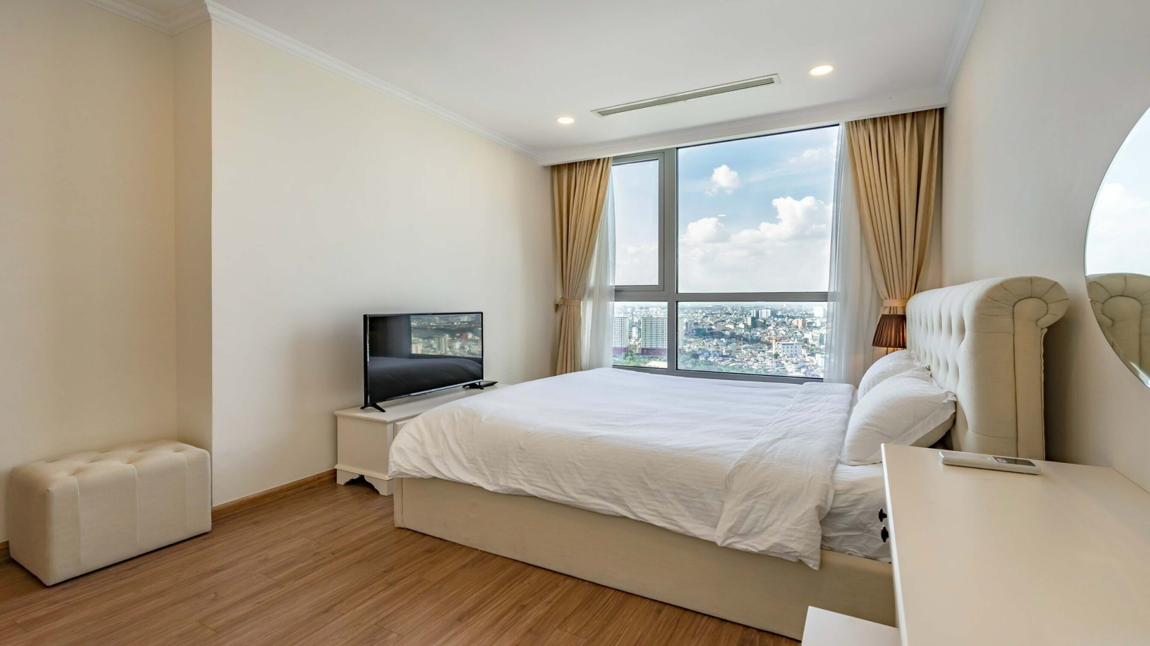 Ein Hotelzimmer mit einem großen Bett, einem Fernseher auf einem Nachttisch, einer kleinen Sitzecke und Blick auf die Stadt durch große Fenster.