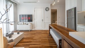 Ein modernes Wohnzimmer mit Holzboden, einem Fernseher, der "NETFLIX" zeigt, einer Küchenzeile mit Kühlschrank und einer Bank.
