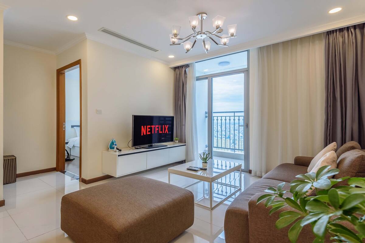 Ein modernes Wohnzimmer mit einem Flachbildfernseher, der 'NETFLIX' anzeigt, einem Sofa, einem Couchtisch und einem Balkon mit Blick auf die Stadt.