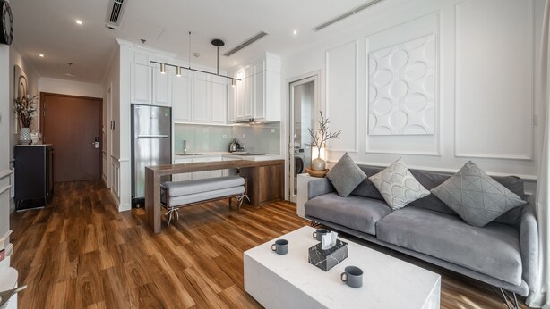 Ein modernes Wohnzimmer mit einem grauen Sofa, einem weißen Couchtisch und einer Küchenzeile mit weißen Schränken und einem Edelstahlkühlschrank.