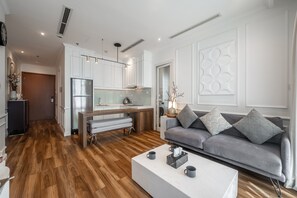 A modern living room with a grey sofa, a white coffee table, and a kitchen area with white cabinets and a stainless steel refrigerator. - Vinhomes Central Park - ANGIA Residence By An Gia Hospitality (Ho Chi Minh City)