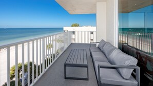 Ein Balkon mit weißer Reling, einer grauen Couch und einem kleinen Tisch. Der Balkon bietet Blick auf einen Strand und den Ozean.