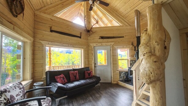 A log cabin interior with a black leather sofa, a patterned armchair, and a stone fireplace. - Forget Me Not Cabin (Sutton-Alpine)