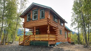 A log cabin with a porch and a staircase, surrounded by trees and mountains. - Forget Me Not Cabin (Sutton-Alpine)