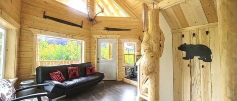 Interior de una cabaña de troncos con techo de madera, un sofá de cuero negro, una silla de madera y la silueta de un oso en la pared.