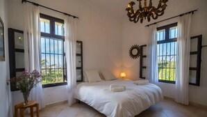 A bedroom with a large bed, two windows with curtains, a chandelier, and a small table with a vase.