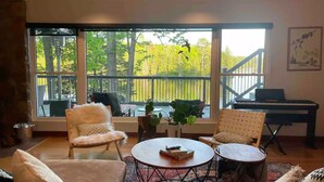 Un salon chaleureux doté d’une grande fenêtre donnant sur une terrasse et des arbres, et aménagé avec un canapé, des fauteuils, une table basse et un piano.