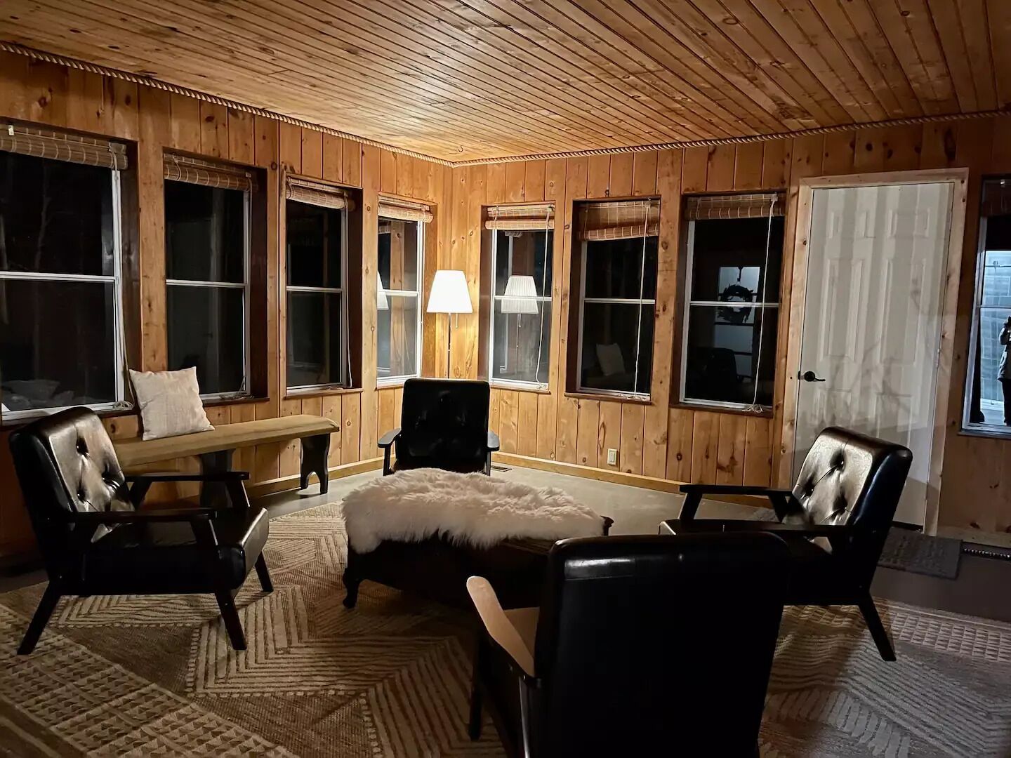 Una habitación acogedora con paredes de madera y ventanas amplias, que incluye un sofá, sillas, un banco y una lámpara.