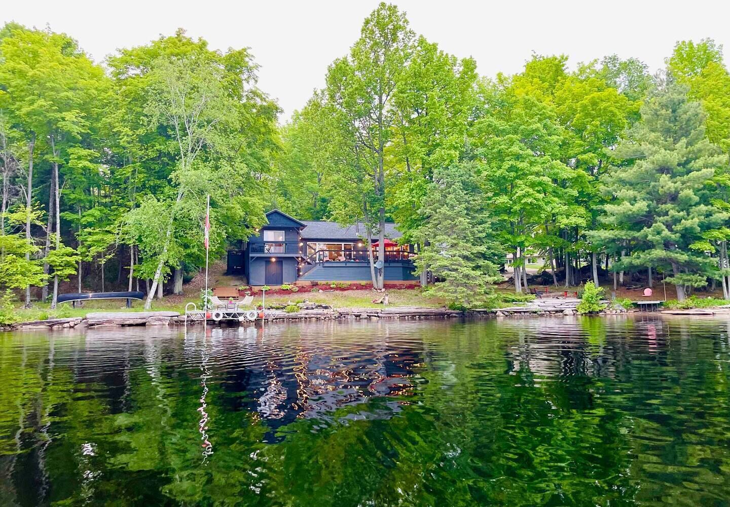 Uma cabana à beira do lago com varanda e área de estar externa, rodeada por uma floresta densa.