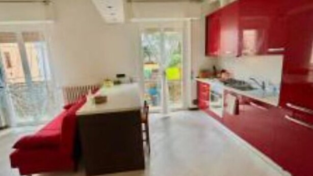 Een moderne keuken met rode kastjes, een zithoek met een rode bank en een groot raam dat zorgt voor daglicht.