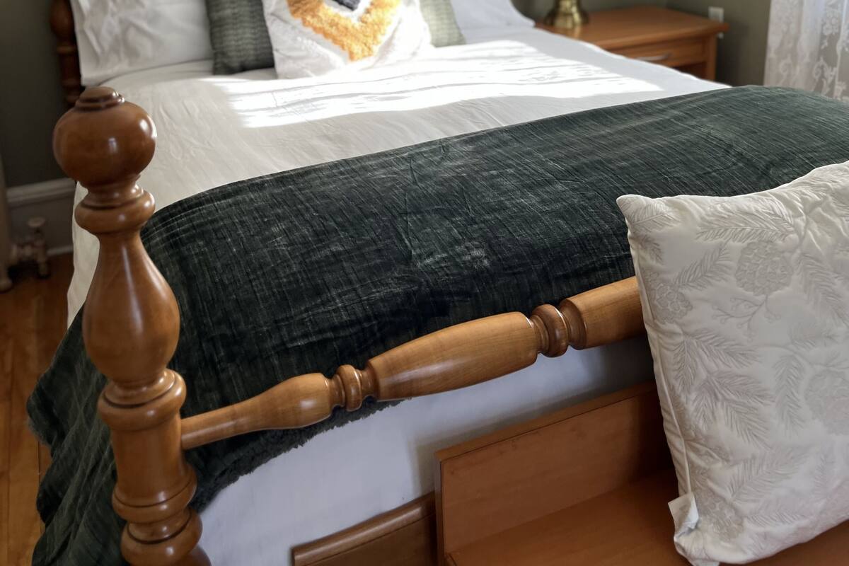 A neatly made bed with a wooden headboard, a green blanket, and multiple pillows. There is a nightstand with a lamp and a framed picture on the wall.