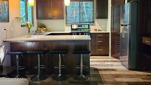 A modern kitchen with a central island, bar stools, wooden cabinets, a stainless steel refrigerator, and pendant lighting.