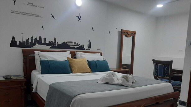 A neatly made bed with a wooden headboard, a bedside table with a lamp, and a wall with a cityscape mural. - LA PROVIDENCIA HOTEL (Neiva)