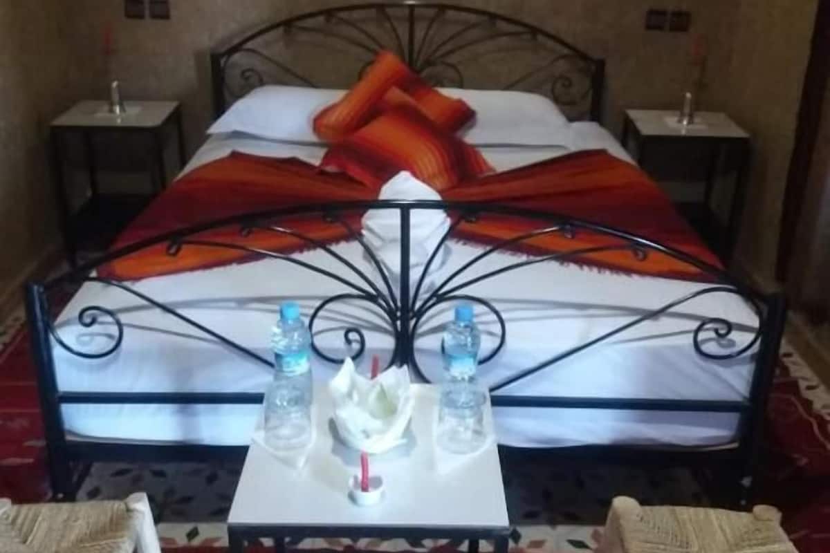 A bed with a metal frame and a colorful blanket, two bedside tables with bottles, a small table with a flower and a drink, and two wicker chairs.