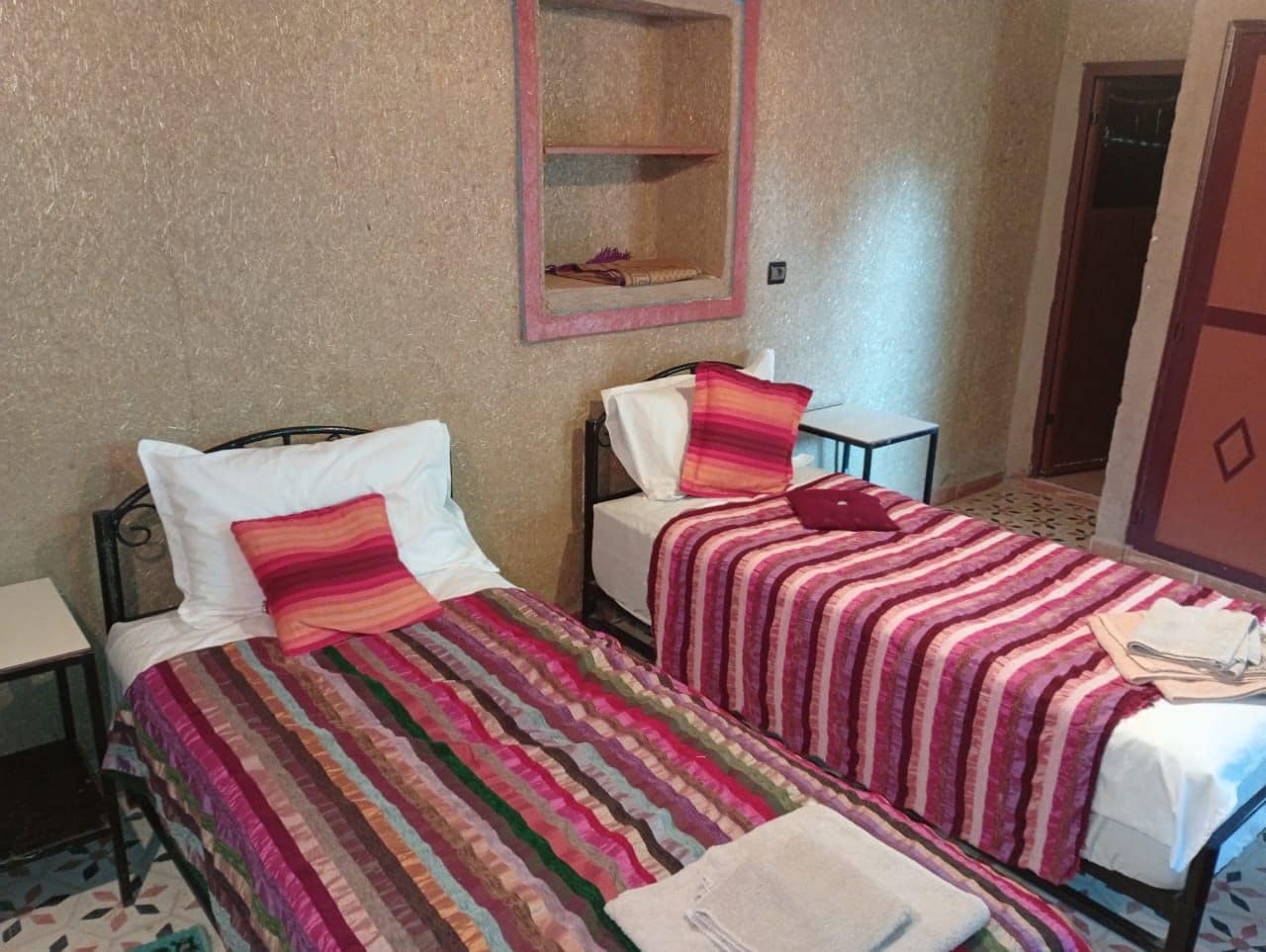 Une chambre avec deux lits, chacun recouvert d’une couvre-lit à rayures et agrémenté d’oreillers, une petite étagère au-dessus des lits et un sol carrelé.