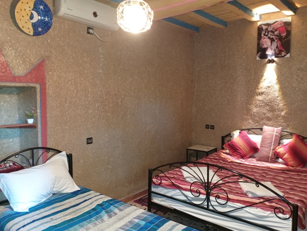 Ein Schlafzimmer mit zwei Betten, eines mit einer gestreiften Tagesdecke und das andere mit einer rot-weißen Musterdecke. Über den Betten befindet sich eine Wandleuchte, und ein kleines Regal mit einem Topfpflanzen.
