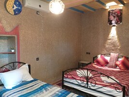 Un dormitorio con dos camas, una con cubrecama a rayas y la otra con un edredón rojo y blanco estampado. Hay una lámpara de pared sobre las camas, y una pequeña estantería con una planta en maceta.