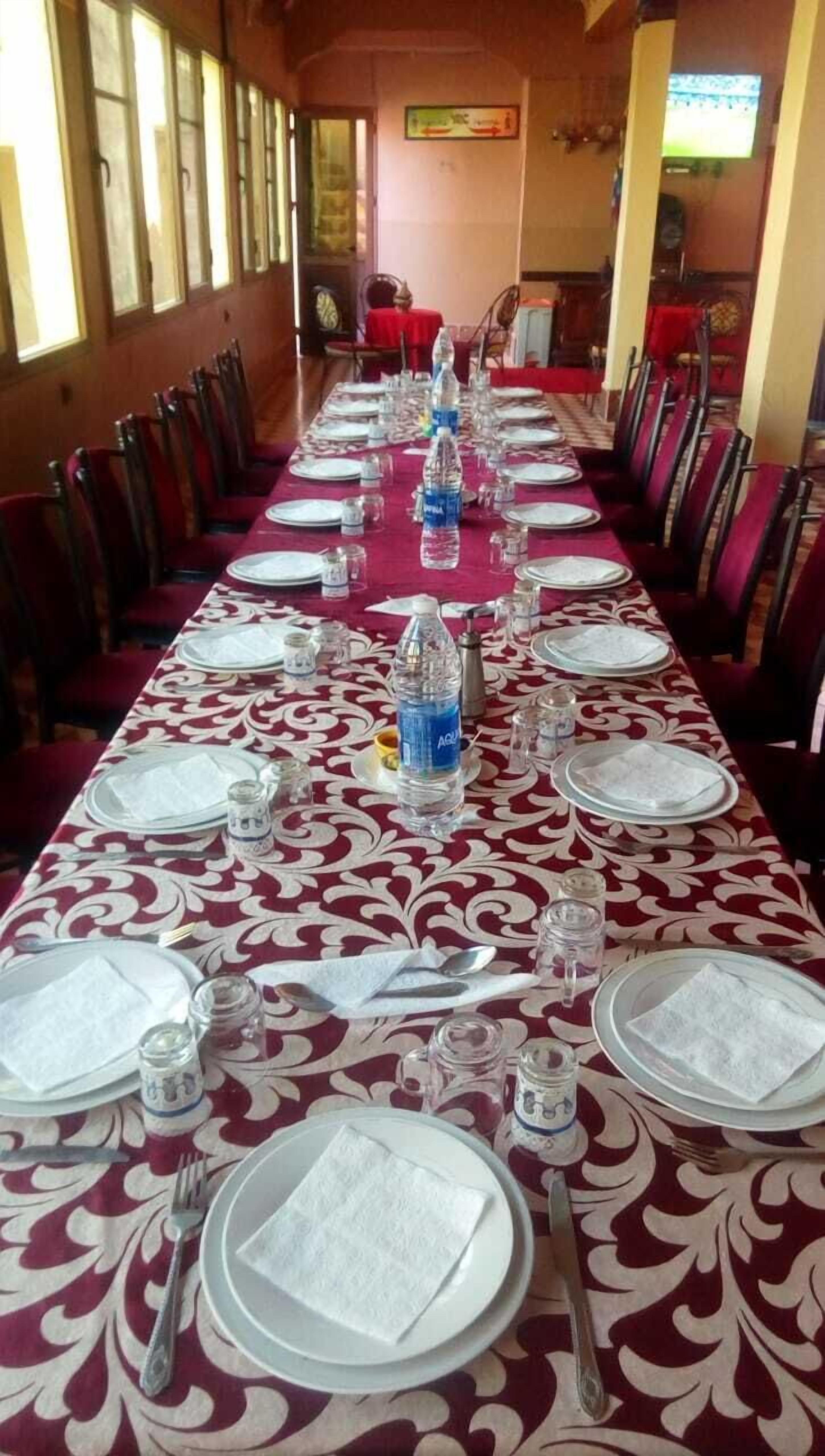 A long dining table set with plates, cutlery, and water bottles, surrounded by chairs in a room with windows and a television.