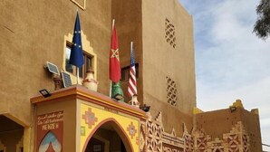 A building with a yellow facade, a decorative entrance with an arch, and flags. - El Menzeh foundation (Rissani)