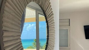 A round mirror with a ribbed frame on a wall, a blue chair, and a window with a view of the ocean.