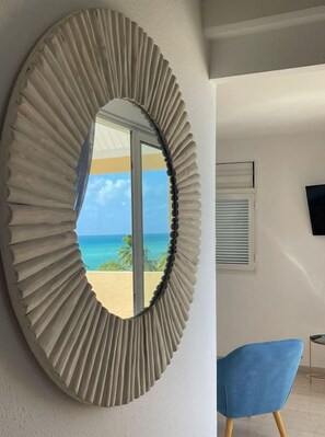 Enjoy a coastal view from the balcony through the large circular mirror, with a blue upholstered chair and flat-screen TV in the modern room.