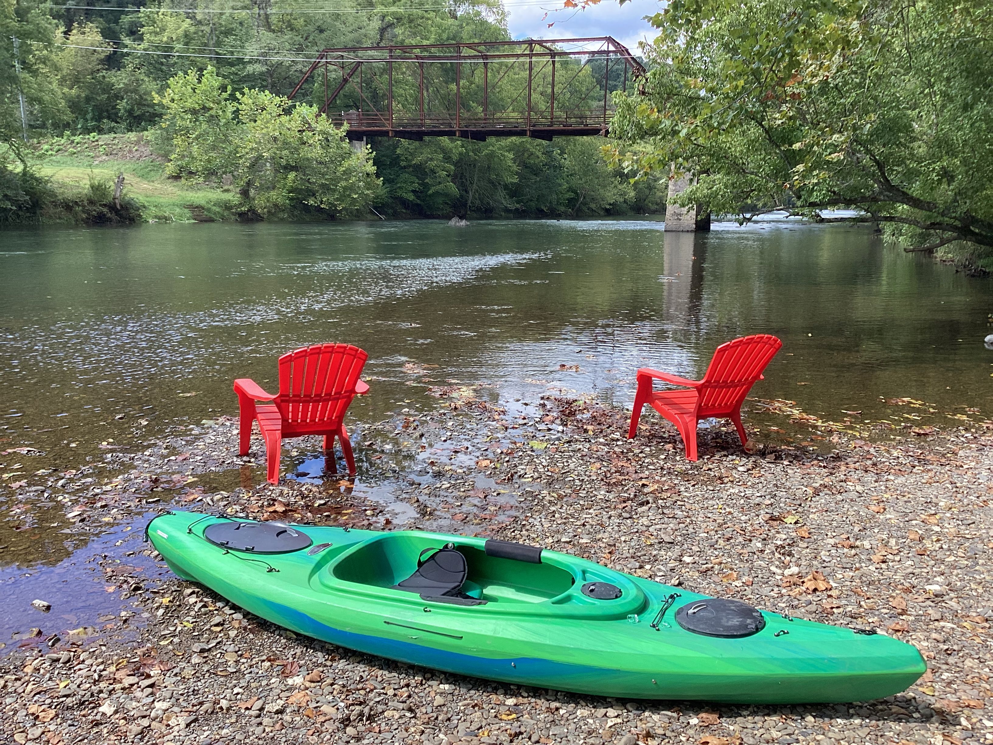Un kayak verde y dos sillas rojas sobre una orilla pedregosa junto a un río con un puente al fondo.
