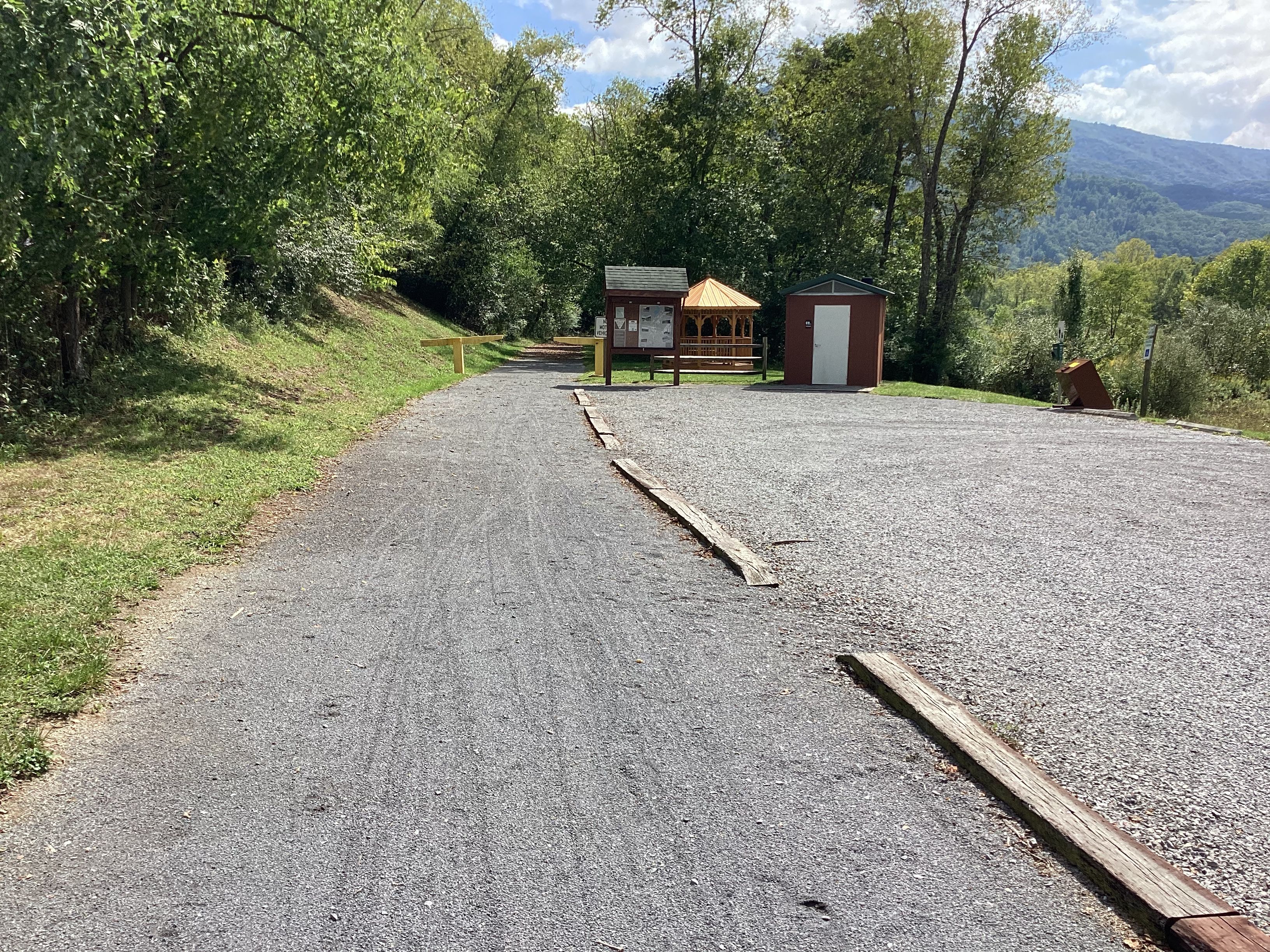 Un chemin de gravier mène à un petit bâtiment sur lequel est apposée une enseigne, le tout étant entouré de verdure et de collines.