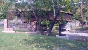 Un puente de piedra rústico con estructura de madera en la parte superior, una rueda metálica grande y un pequeño edificio de ladrillo al fondo.