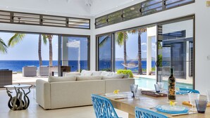 A modern living room with a large sofa, dining table, and chairs, overlooking a pool and the ocean.